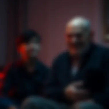 A young man looks uneasy while his dad sits silently with a strange smile in a dimly lit room.