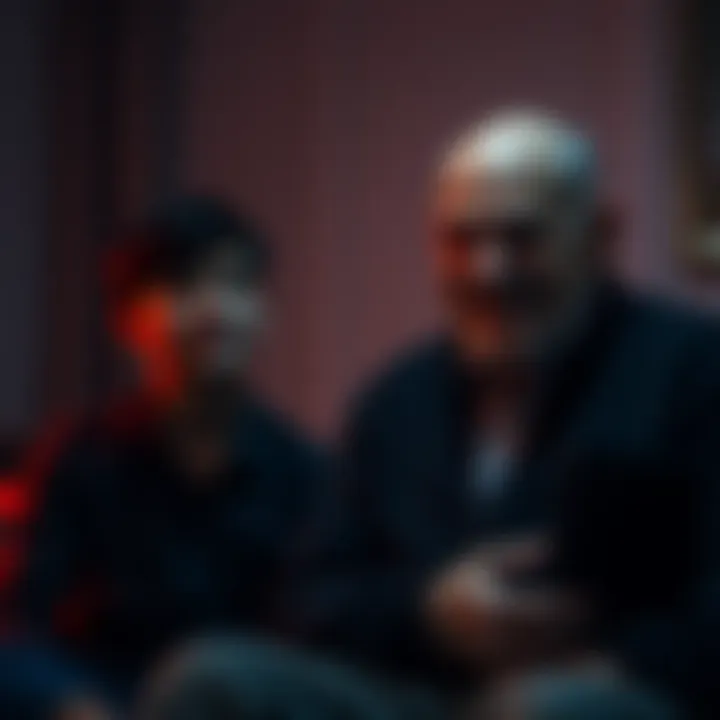 A young man looks uneasy while his dad sits silently with a strange smile in a dimly lit room.